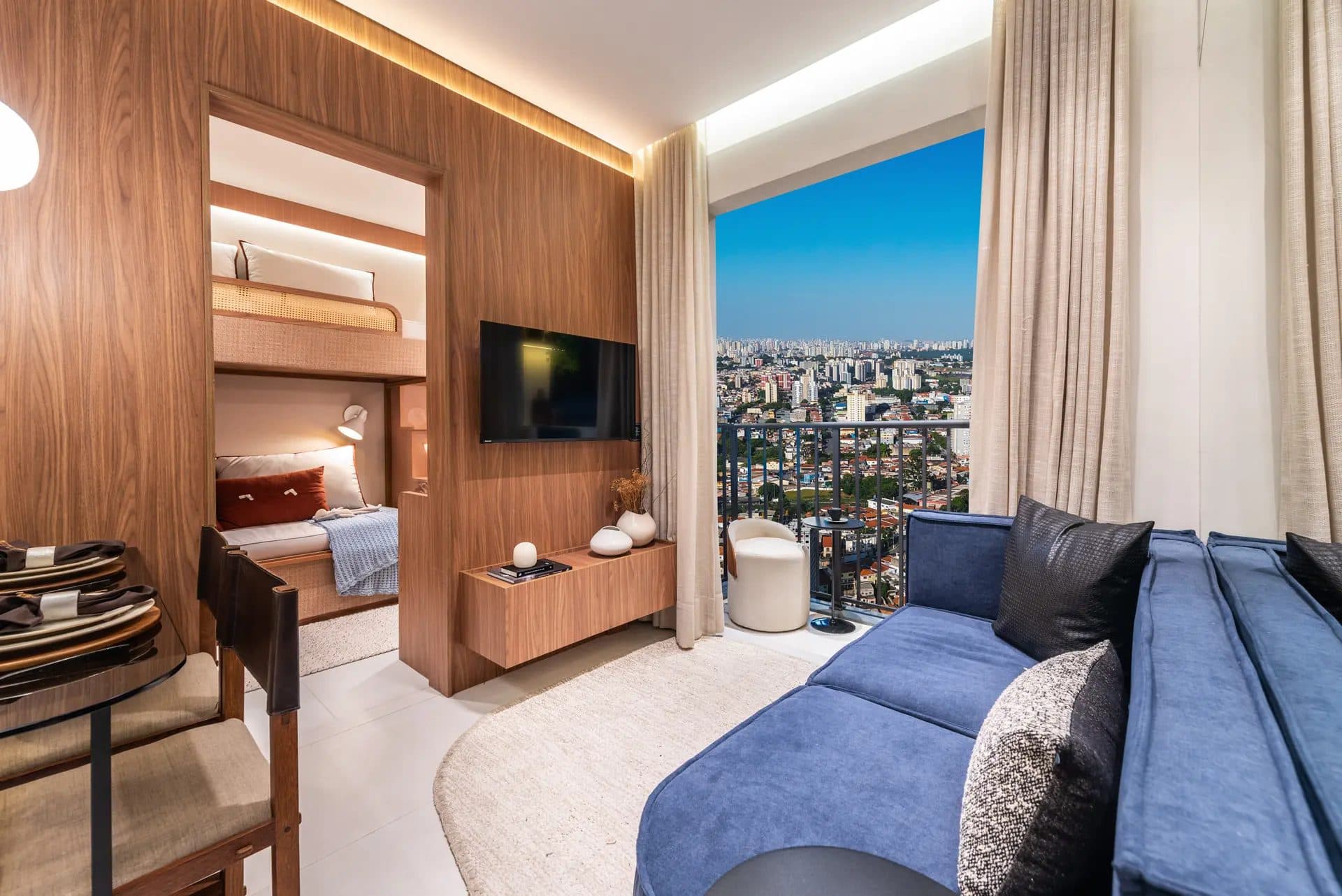 Sala de estar com painel de madeira para TV, sofá azul e varanda com vista para a cidade.